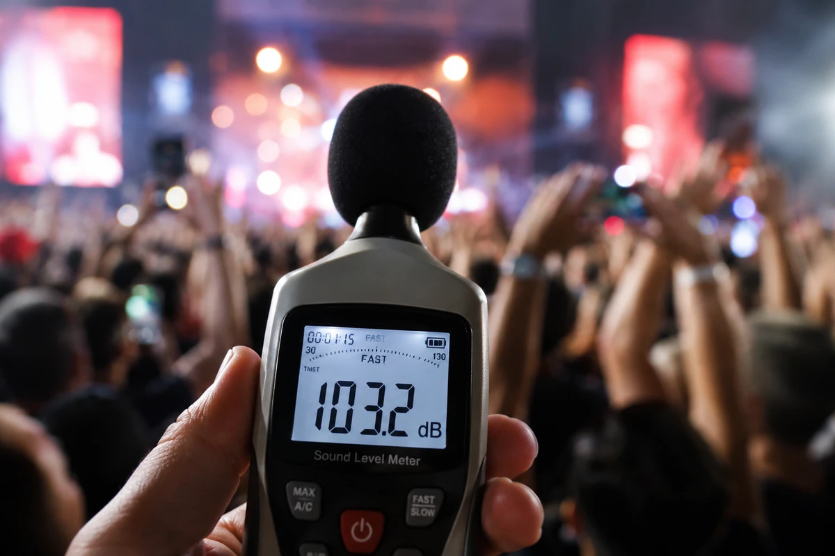 Decibel Là Gì Và Ngưỡng Âm Thanh An Toàn Cho Tai Người 7 Cận cảnh máy đo decibel hiển thị mức âm thanh hơn 100dB tại khu vực khán giả trong sự kiện âm nhạc, phía sau là sân khấu và đám đông đang cổ vũ sôi động